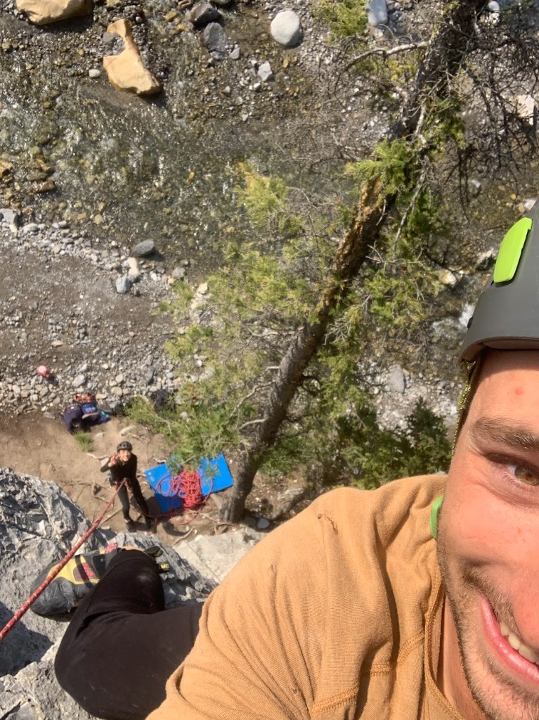 Rock climbing selfie from high on a cliff, rope and partner visible far below